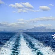 Blick von einer Fähre auf griechische Inseln in der Ferne, mit einer im Wind wehenden griechischen Flagge im Vordergrund