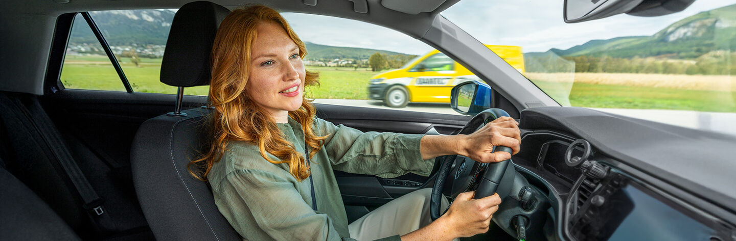 Eine rothaarige Frau mit grüner Bluse sitzt in einem Auto und hält mit beiden Händen das Lenkrad.