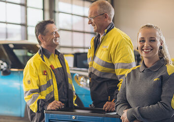 Zwei männliche und eine weibliche Kfz-Technikerin stehen an einer Werkbank in der Kfz-Technik Werkstatt. Im Hintergrund ein blaues Auto auf der Hebebühne. Ein weiterer Kollege kommt im Hintergrund auf die drei Personen zu.