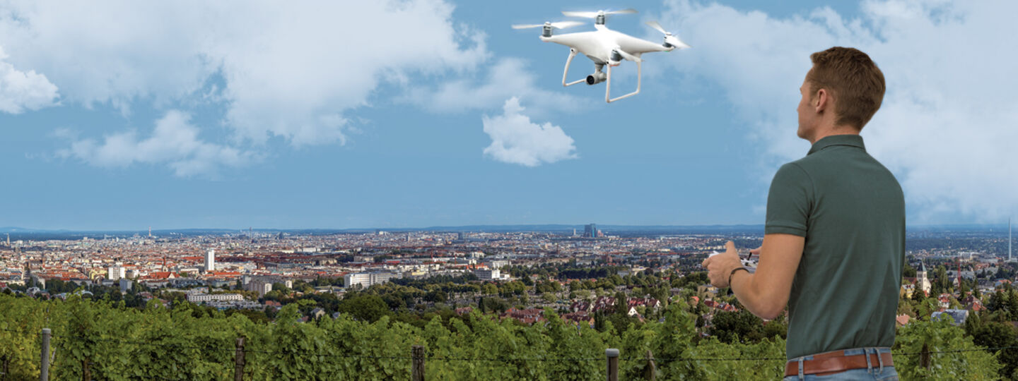 Ein Mann steuert eine weiße Drohne, die vor ihm in der Luft fliegt. Er steht auf einem Aussichtplatz am Rande einer Großstadt.
