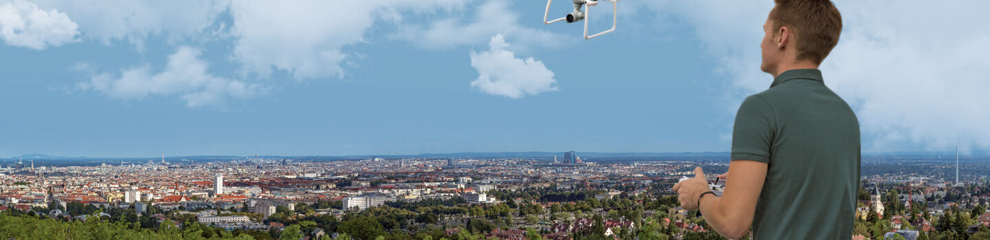 Ein Mann steuert eine weiße Drohne, die vor ihm in der Luft fliegt. Er steht auf einem Aussichtplatz am Rande einer Großstadt.