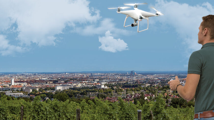 Ein Mann steuert eine weiße Drohne, die vor ihm in der Luft fliegt. Er steht auf einem Aussichtplatz am Rande einer Großstadt.