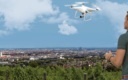 Ein Mann steuert eine weiße Drohne, die vor ihm in der Luft fliegt. Er steht auf einem Aussichtplatz am Rande einer Großstadt.