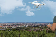 Ein Mann steuert eine weiße Drohne, die vor ihm in der Luft fliegt. Er steht auf einem Aussichtplatz am Rande einer Großstadt.