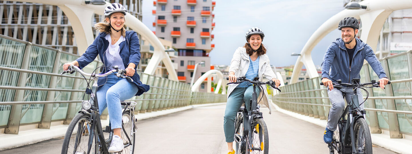 Zwei junge Frauen und ein Mann fahren auf einer Fahrradbrücke in einem Wohnviertel. Alle drei tragen Fahrradhelme.