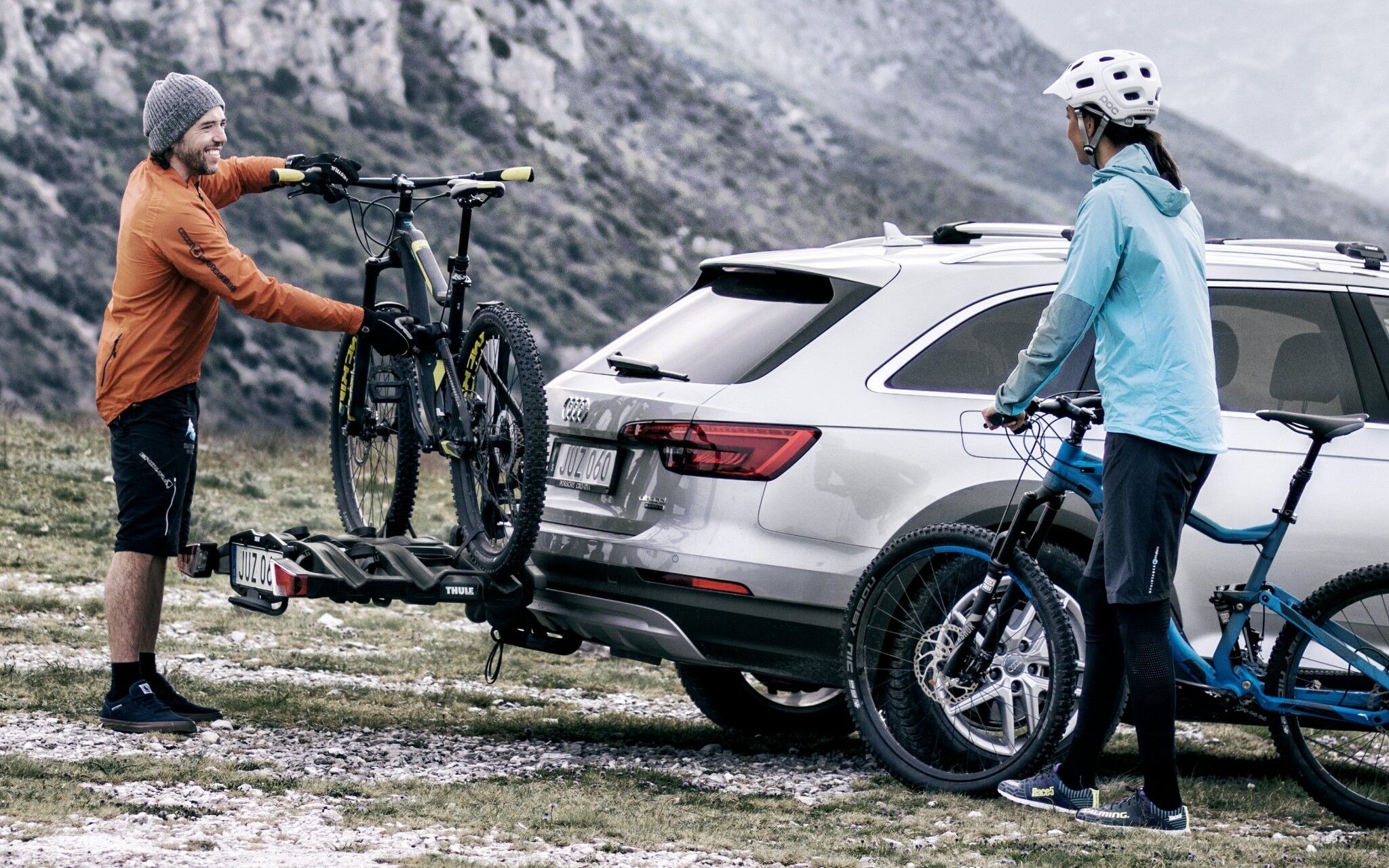 Ein Mann stellt auf einen Thule-Heckträger am Auto ein Mountain-Bike, neben dem Auto steht eine Frau und hält ein Fahrrad in der Hand.