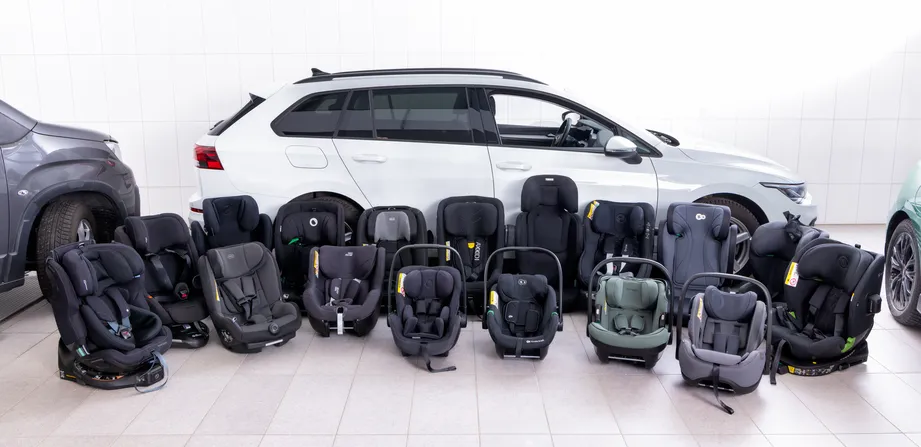 16 Kindersitze stehen nebeneinander am Boden vor einem weißen Auto 
