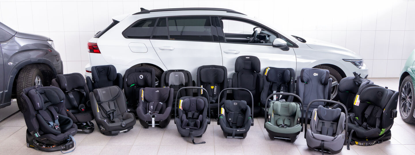 16 Kindersitze stehen nebeneinander am Boden vor einem weißen Auto 