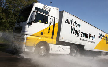 Ein weiß-gelber LKW mit ÖAMTC Fahrtechnik-Beschriftung fährt nach links. Wasser spritzt davon. Im Hintergrund blauer Himmel und Bäume.