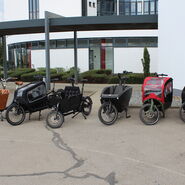 5 einspurige E-Lastenfahrräder stehen vor Haus.