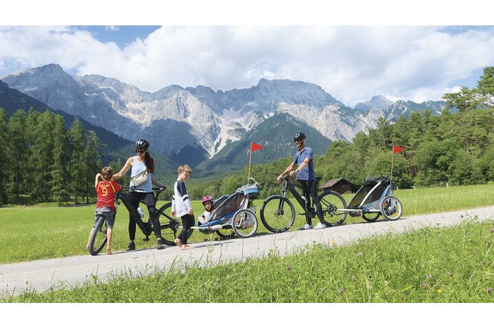 Eine Familie mit 3 Kleinkindern ist mit 2 Radanhängern unterwegs. Im Hintergrund die Bergkulisse.
