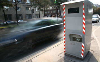 Rechts im Bild sieht man eine Radarbox, bei der ein schwarzes Auto auf der Straße vorbeifährt.