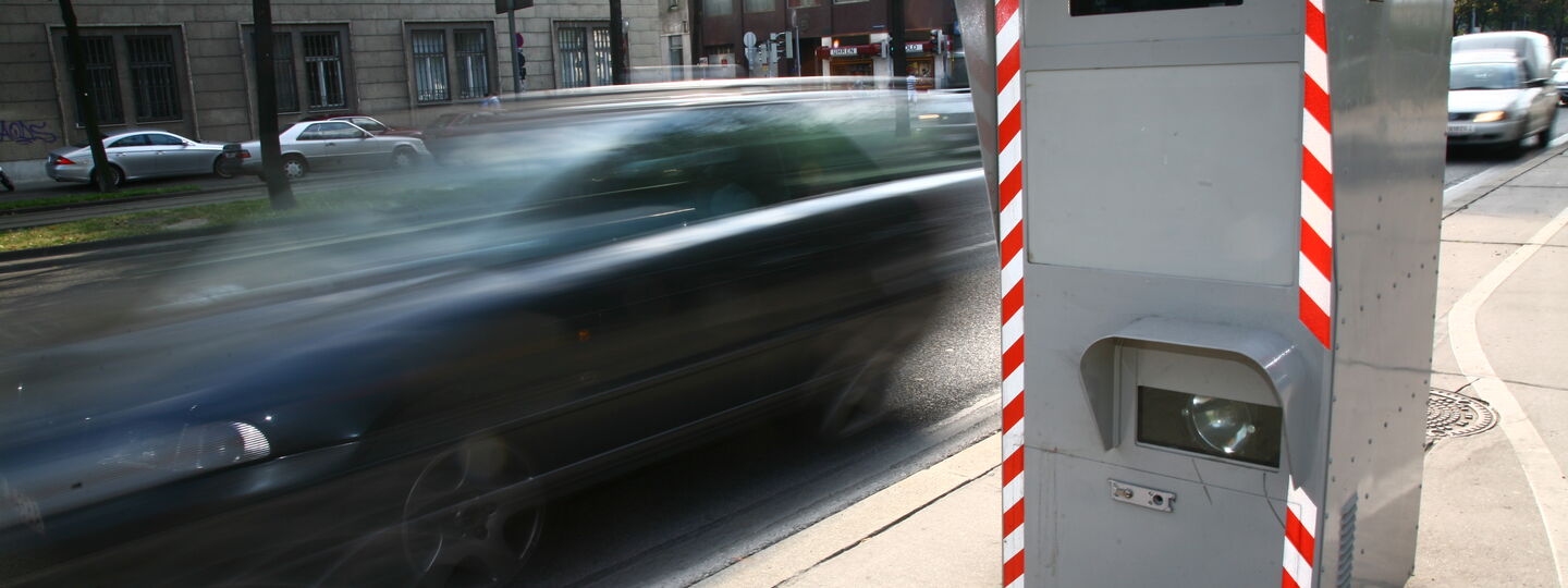 Rechts im Bild sieht man eine Radarbox, bei der ein schwarzes Auto auf der Straße vorbeifährt.