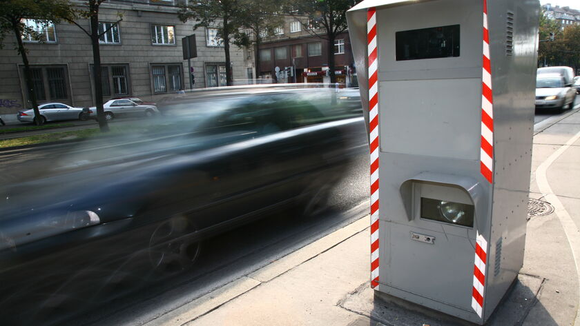 Rechts im Bild sieht man eine Radarbox, bei der ein schwarzes Auto auf der Straße vorbeifährt.