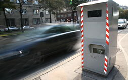 Rechts im Bild sieht man eine Radarbox, bei der ein schwarzes Auto auf der Straße vorbeifährt.