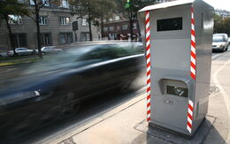 Rechts im Bild sieht man eine Radarbox, bei der ein schwarzes Auto auf der Straße vorbeifährt.