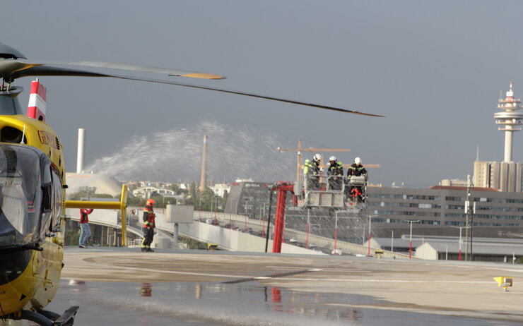 Das Bild zeigt eine Übungssituation auf dem Hubschrauberlandeplatz am Heliport des ÖAMTC-Mobilitätszentrums in Wien-Erdberg. Links im Bild steht ein Christophorus 9-Rettungshubschrauber, während im Hintergrund mehrere Feuerwehrleute auf einer Teleskopmastbühne stehen. Sie tragen Schutzhelme und Ausrüstung.