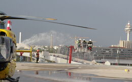 Das Bild zeigt eine Übungssituation auf dem Hubschrauberlandeplatz am Heliport des ÖAMTC-Mobilitätszentrums in Wien-Erdberg. Links im Bild steht ein Christophorus 9-Rettungshubschrauber, während im Hintergrund mehrere Feuerwehrleute auf einer Teleskopmastbühne stehen. Sie tragen Schutzhelme und Ausrüstung.