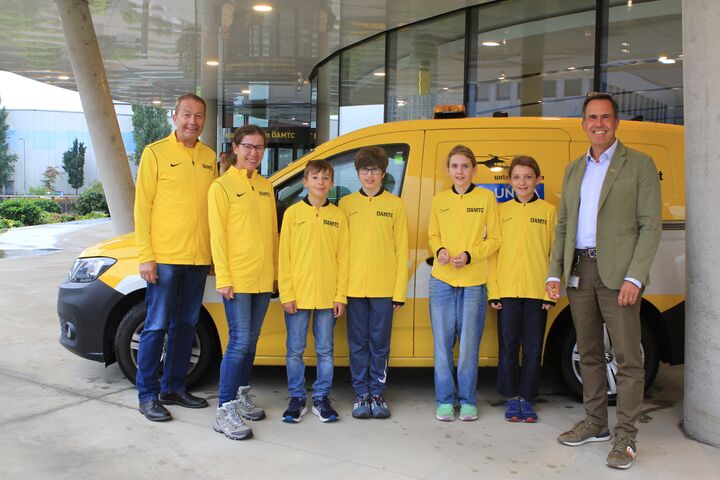 Vier Kinder in gelben ÖAMTC-Trikots stehen neben drei Erwachsenen vor einem ÖAMTC Pannenfahrzeug