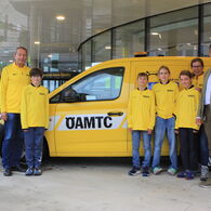 Vier Kinder in gelben ÖAMTC-Trikots stehen neben vier Erwachsenen vor einem ÖAMTC Pannenfahrzeug