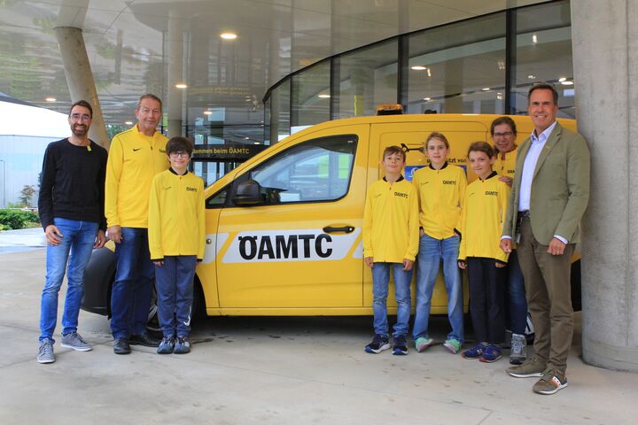 Vier Kinder in gelben ÖAMTC-Trikots stehen neben vier Erwachsenen vor einem ÖAMTC Pannenfahrzeug