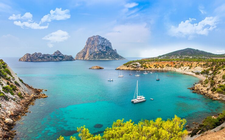 Ibiza Blick auf eine traumhafte Bucht auf Ibiza mit türkisblauem Wasser, Segelschiffen und der Felseninsel Es Vedrà im Hintergrund.