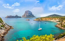 Blick auf eine traumhafte Bucht auf Ibiza mit türkisblauem Wasser, Segelschiffen und der Felseninsel Es Vedrà im Hintergrund.