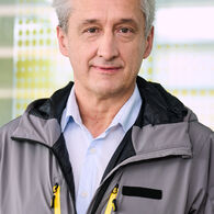 KERBL Steffan (2) Das Foto zeigt einen Mann in einer grauen Jacke, unter der er ein hellblaues Hemd trägt. Der Mann hat graue Haare und steht vor einer Glaswand mit gelben Punkten.