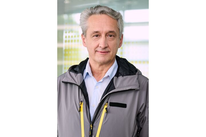 Das Foto zeigt einen Mann in einer grauen Jacke, unter der er ein hellblaues Hemd trägt. Der Mann hat graue Haare und steht vor einer Glaswand mit gelben Punkten.