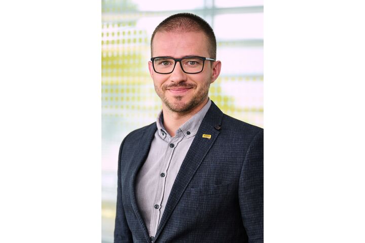 Ein Mann mit kurzen Haaren, Brille und Bart. Er trägt ein dunkles Sakko über einem grauen Hemd.