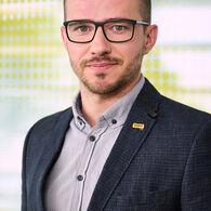 Ein Mann mit kurzen, dunklen Haaren und schwarz umrandeter Brille. Er trägt ein dunkelblaues Sakko, darunter ein Helles Hemd.