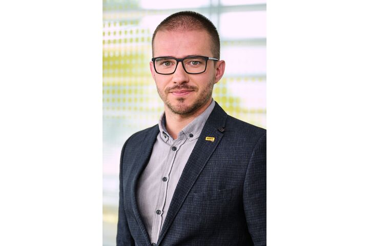 Ein Mann mit kurzen, dunklen Haaren und schwarz umrandeter Brille. Er trägt ein dunkelblaues Sakko, darunter ein Helles Hemd.