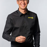 Porträt eines lächelnden jungen Mannes mit Brille und kurzen Haaren, in Schwarz gekleidet. Auf dem Hemd sieht man den gelben Aufdruck "ÖAMTC Fahrtechnik"