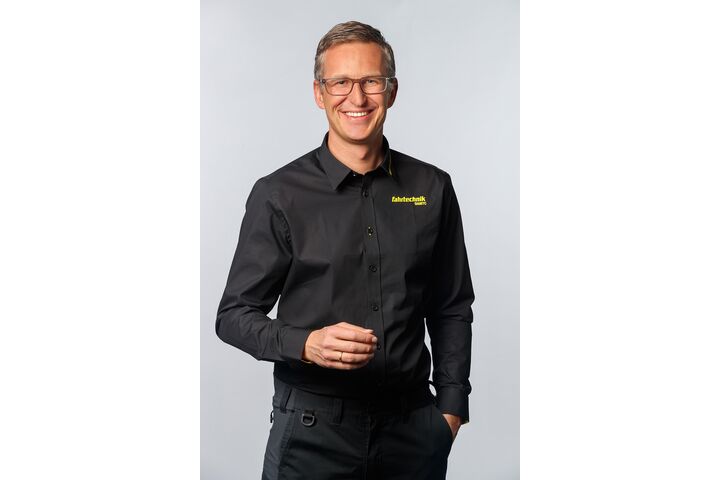 Porträt eines lächelnden jungen Mannes mit Brille und kurzen Haaren, in Schwarz gekleidet. Auf dem Hemd sieht man den gelben Aufdruck "ÖAMTC Fahrtechnik"