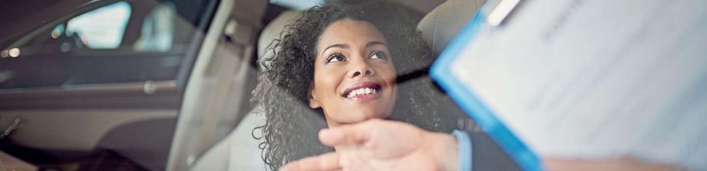 Lächelnde, junge Frau sitzt in einem Auto auf dem Fahrersitz während ein Mann mit einem Klemmbrett ihr etwas erklärt.