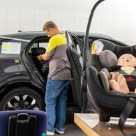 Kindersitztest Herbst 2025 - 2 Im Vordergrund liegt eine Puppe in einem Kindersitz, im Hintergrund bereitet ein Mann in ÖAMTC-Kleidung alles für den Test im Auto vor.