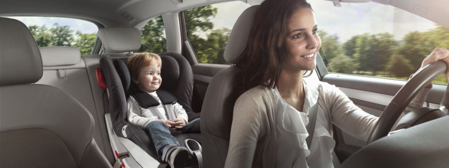 Eine Frau sitzt am Fahrersitz und fährt lächelnd ein Auto. Auf der Rückbank ist ein Kleinkind im Kindersitz.
