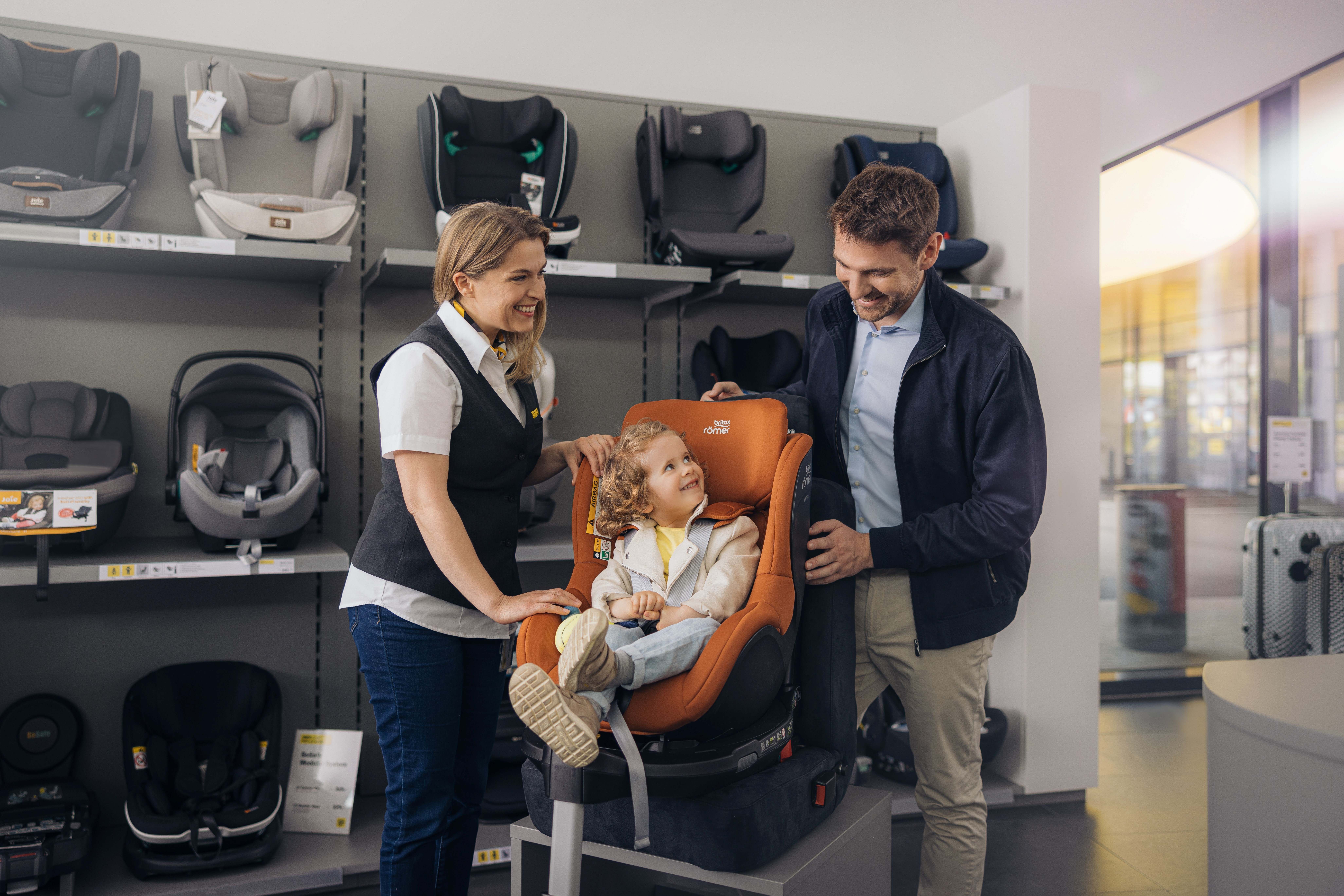 Eine Dame berät einen Herren zum Thema Kindersitzkauf. In der Mitte sitzt ein Kleinkind in einem Kindersitz.