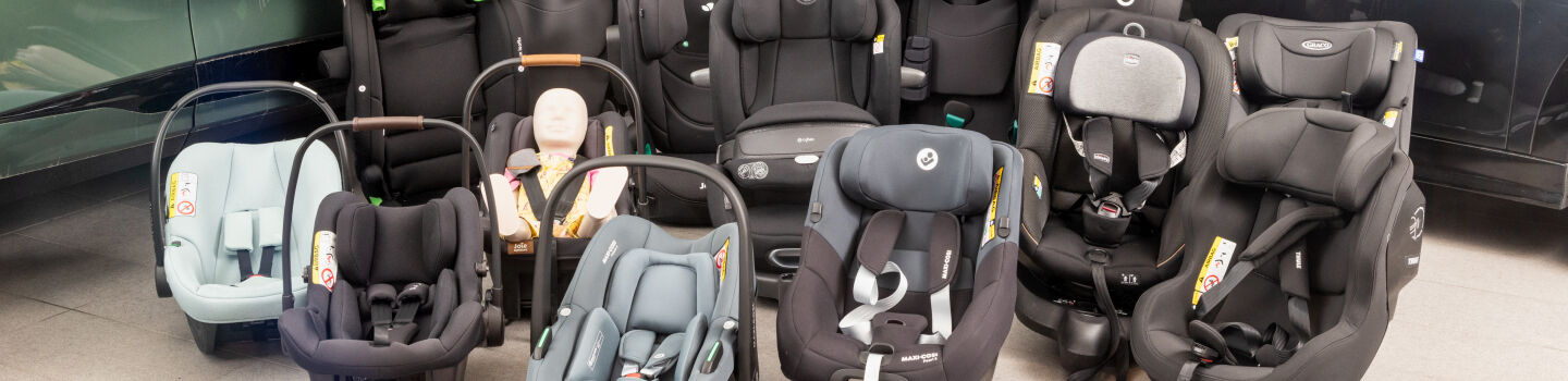 16 Kindersitze in unterschiedlichen Größen und Farben stehen vor zwei Autos am Boden einer Testhalle.