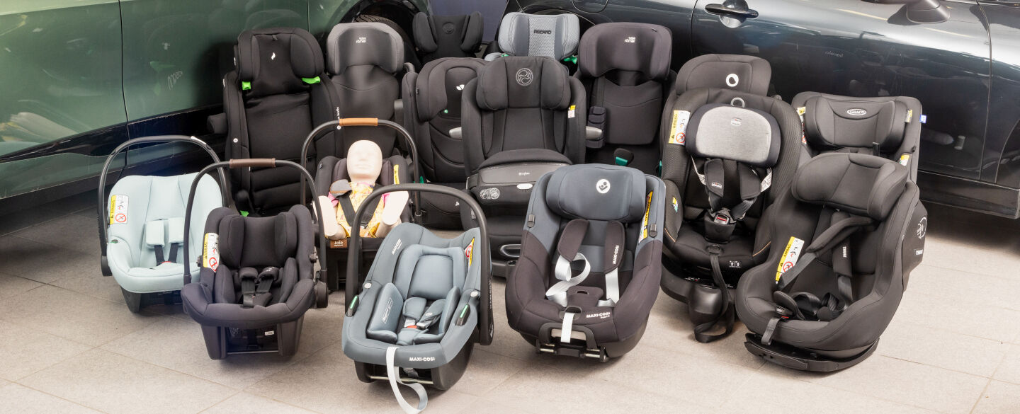 Kindersitztest 2025.jpg 16 Kindersitze in unterschiedlichen Größen und Farben stehen vor zwei Autos am Boden einer Testhalle.