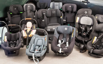 16 Kindersitze in unterschiedlichen Größen und Farben stehen vor zwei Autos am Boden einer Testhalle.