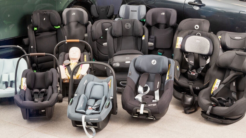 16 Kindersitze in unterschiedlichen Größen und Farben stehen vor zwei Autos am Boden einer Testhalle.