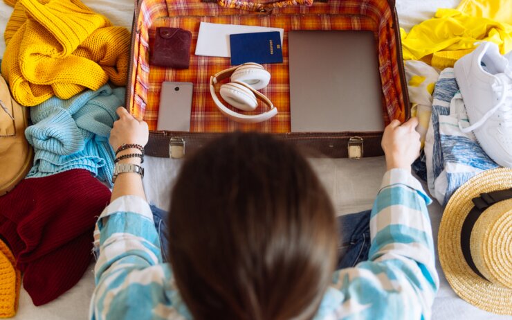 Eine Person ist gerade dabei ihre Kleidung, Schuhe und elektronische Geräte in einen offenen Koffer auf einem Bett zu packen.