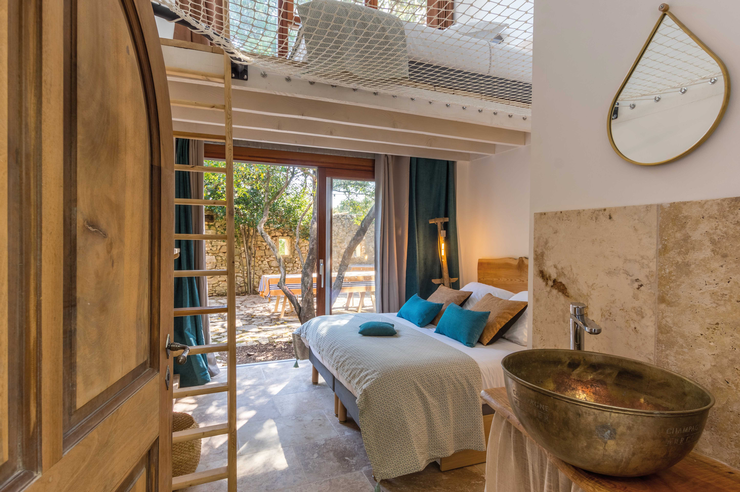 Ein Zimmer mit einem normalen Bett und einem Netzbett an der Decke, Holztüren, einem großen Waschbecken und Blick auf den Garten.