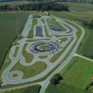 Inmitten einer ländlichen Landschaft erstreckt sich das Fahrtechnik Zentrum Lang/Lebring. Es gibt viele asphaltierte Trainingsstrecken mit Kreuzungselementen, Kreisbahn und Kurvenkombinationen. Im Hintergrund ist ein Gebäude.