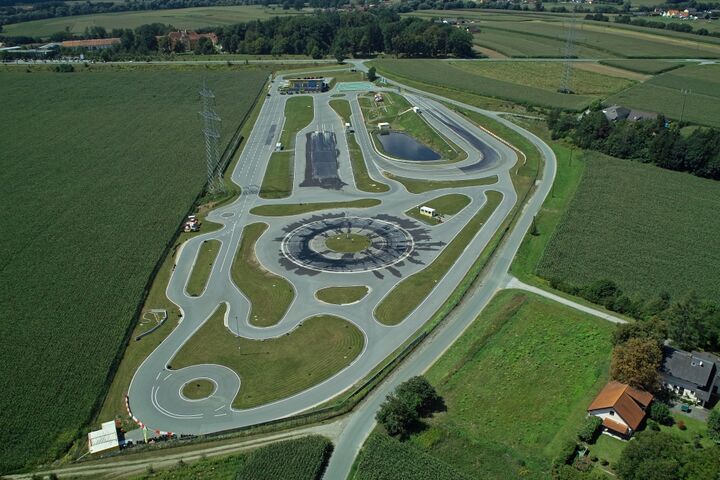 Inmitten einer ländlichen Landschaft erstreckt sich das Fahrtechnik Zentrum Lang/Lebring. Es gibt viele asphaltierte Trainingsstrecken mit Kreuzungselementen, Kreisbahn und Kurvenkombinationen. Im Hintergrund ist ein Gebäude.