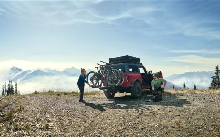 Ein Fahrzeug vor einer Bergkulisse mit zwei Personen. Am Heck des Autos ein Thule-Heckträger, auf welchem zwei Fahrräder befestigt sind. 
