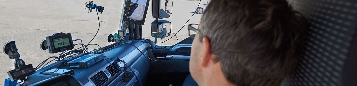 Lkw-Fahrer blickt nach rechts in den Seitenspiegel, wo ein Fußgänger sichtbar ist. Am Armaturenbrett befinden sich verschiedene Assistenzsysteme.