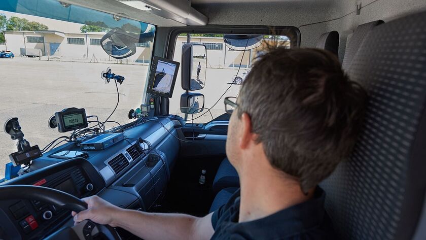 Lkw-Fahrer blickt nach rechts in den Seitenspiegel, wo ein Fußgänger sichtbar ist. Am Armaturenbrett befinden sich verschiedene Assistenzsysteme.