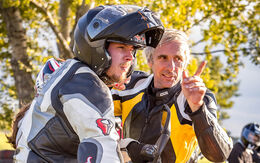 Ein Mann in Motorradkleidung und Helm steht neben einem Instruktor. Der Instruktor zeigt mit dem Finger in eine Richtung und erklärt etwas. Im Hintergrund sind Bäume.